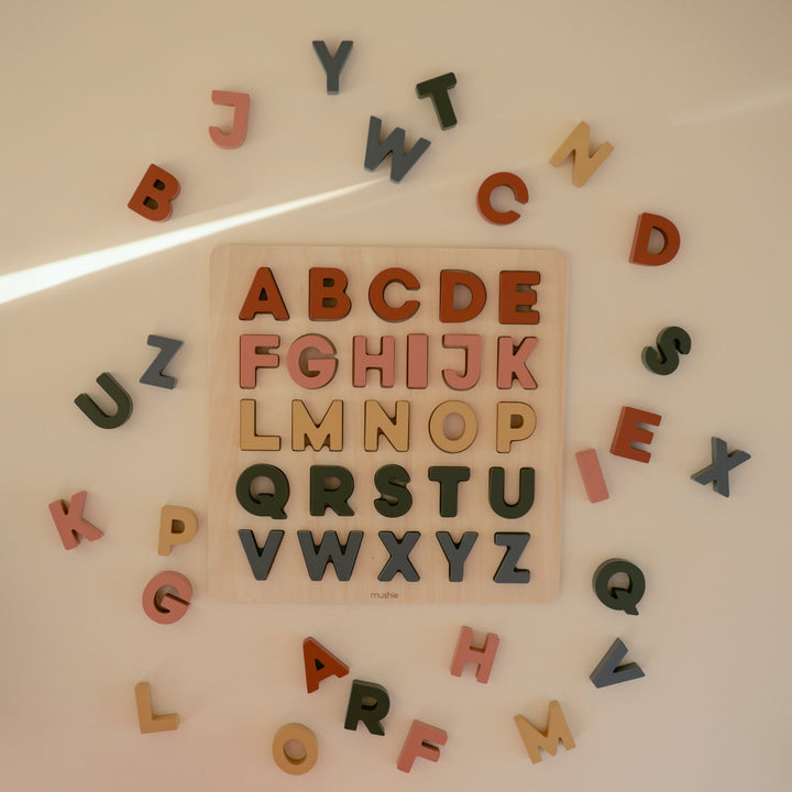 A top-down view of a square wooden alphabet puzzle surrounded by scattered loose alphabet pieces on a light surface, with the puzzle board displaying letters from A to Z.