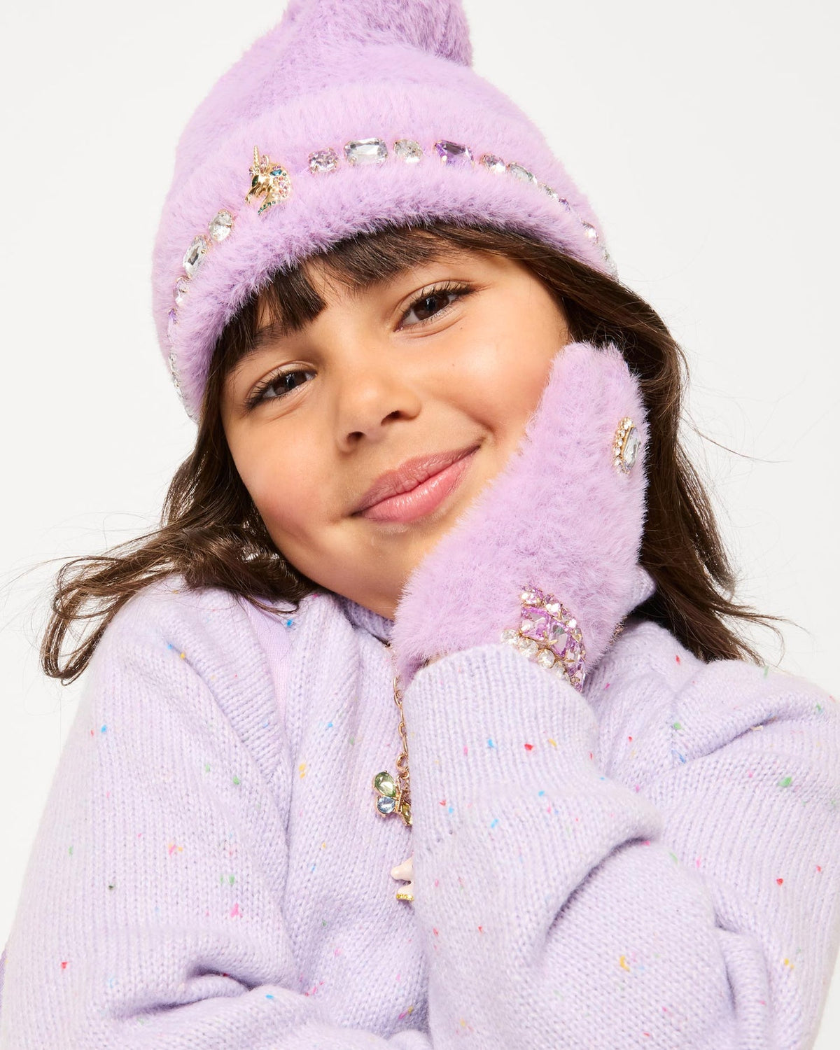 A young girl wearing a fuzzy lavender knit beanie and matching knit mittens with jeweled embellishments, posed against a clean white background.