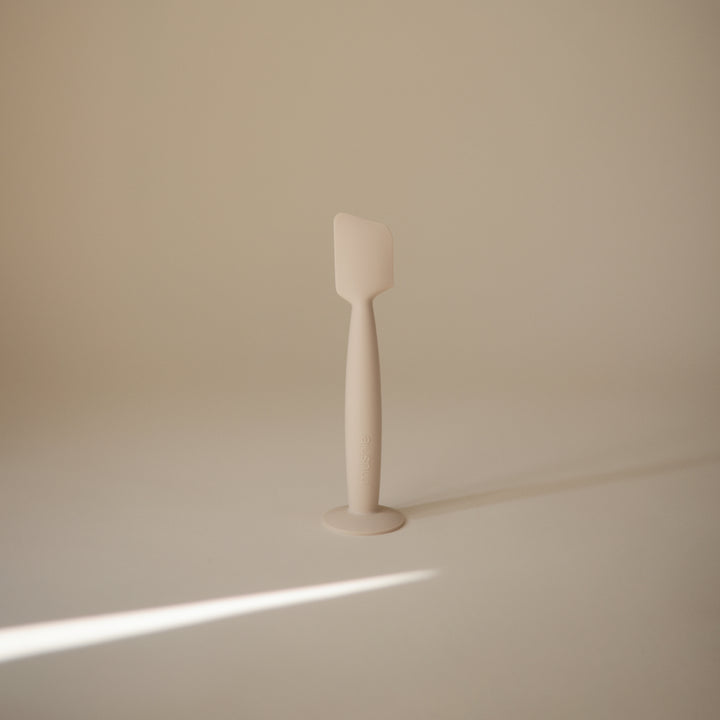 A minimalist, matte off-white silicone spatula standing upright on a matching circular base against a solid beige background.