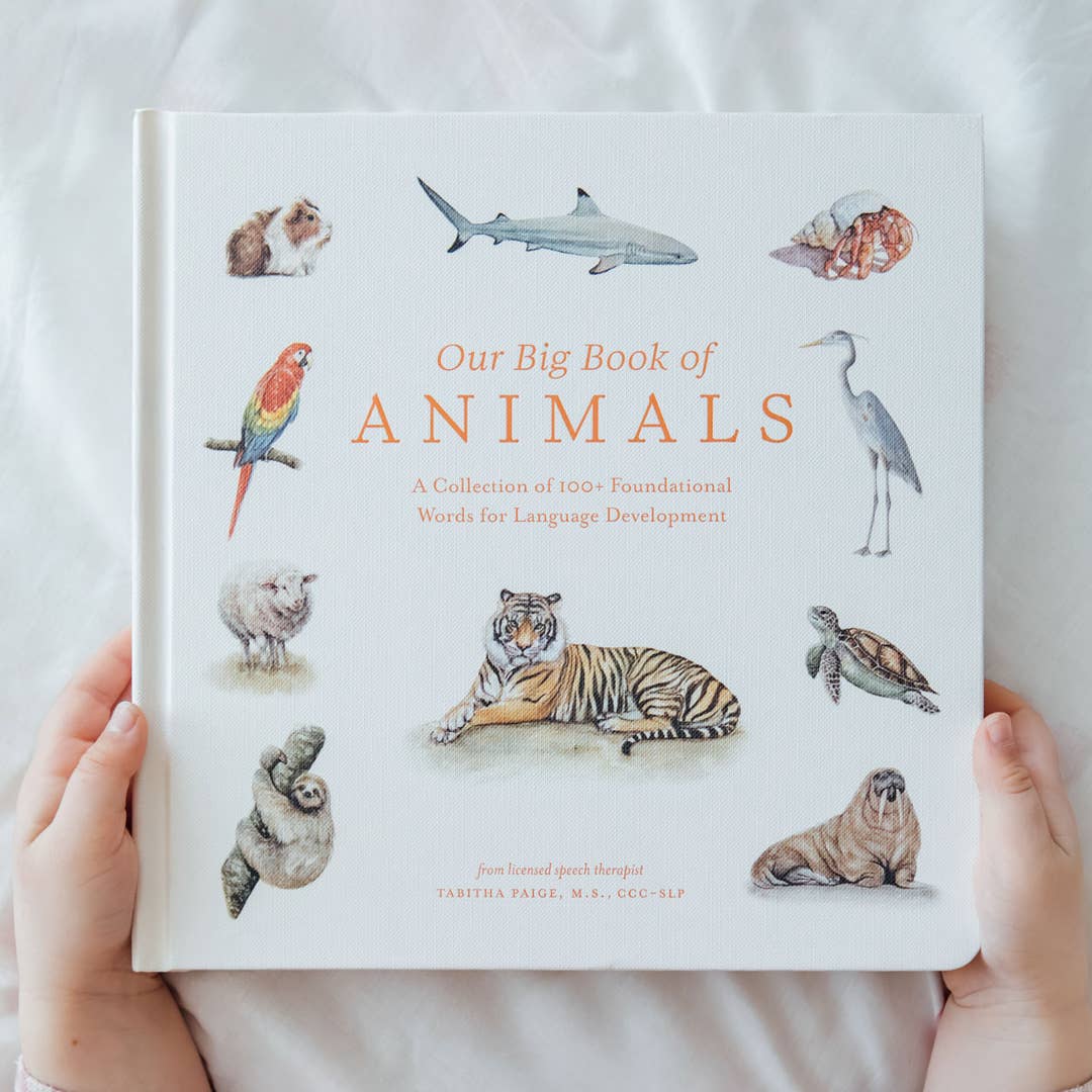 A person holds a hardcover children's book titled "Our Big Book of Animals" against a soft white fabric background. The cover features watercolor-style illustrations of various animals, including a shark, parrot, turtle, tiger, and sloth, arranged around the title.