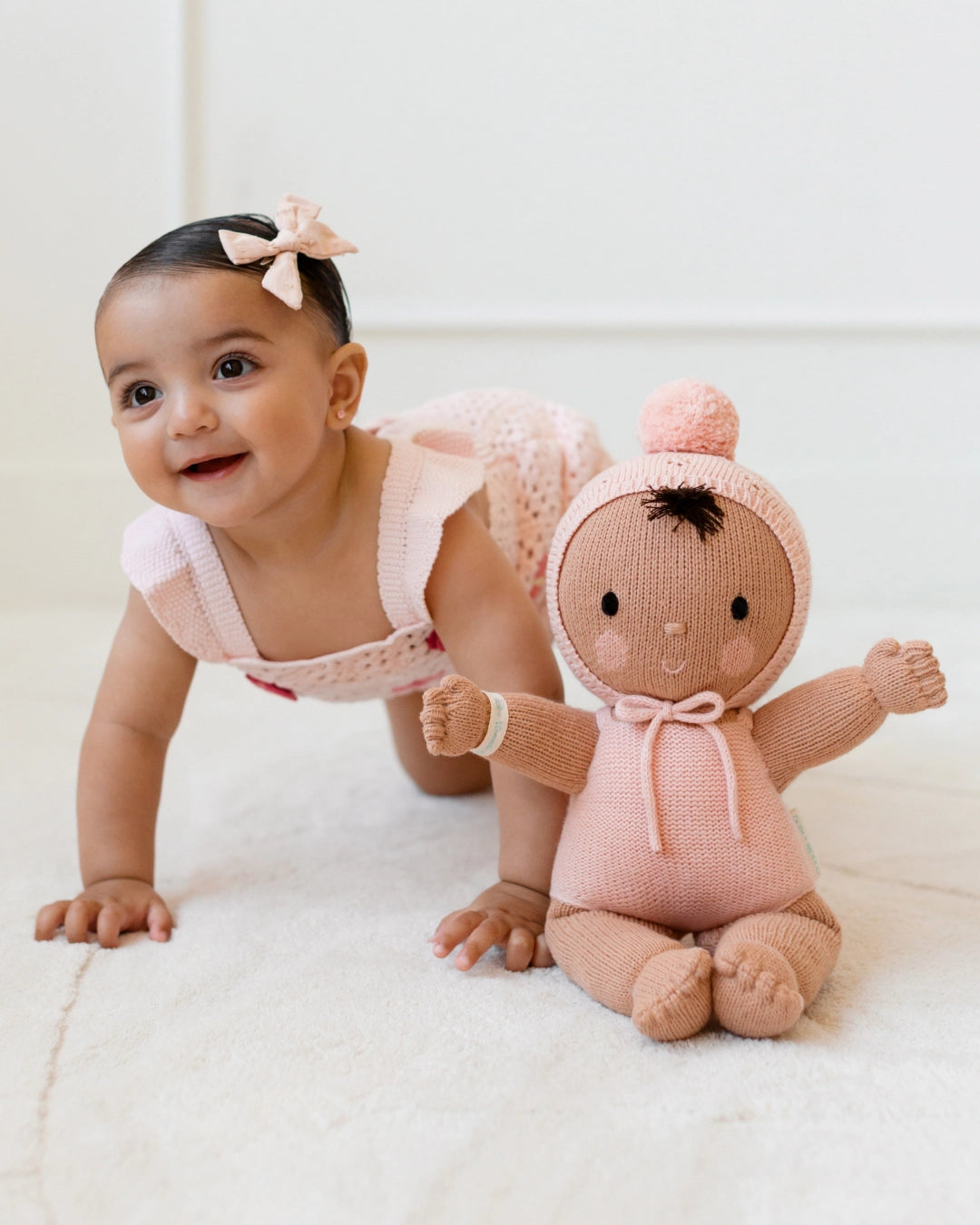 Baby crawling next to a knitted doll wearing a pink outfit on a white background