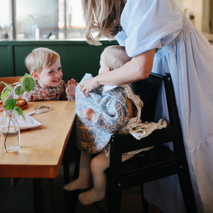 Woman helping a child sit on a chair at a table with another child watching.