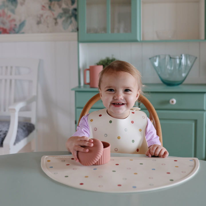 Child sitting at a table with a polka dot placemat and bib, holding a pink bowl.
