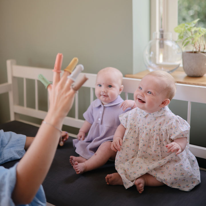 Two babies sitting on a bed with a person's hand reaching out, likely playing with them.