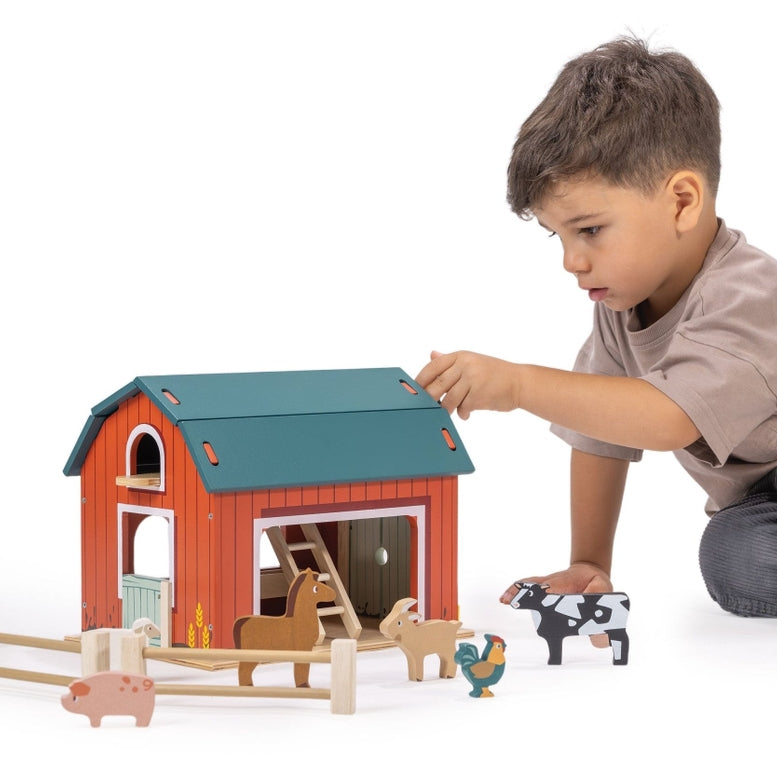 A young boy with short brown hair, wearing a gray t-shirt and jeans, plays with the Wooden Big Red Barn Playset by Mentari, reaching for the roof of the red barn while wooden farm animal figures stand in the fenced enclosure below.