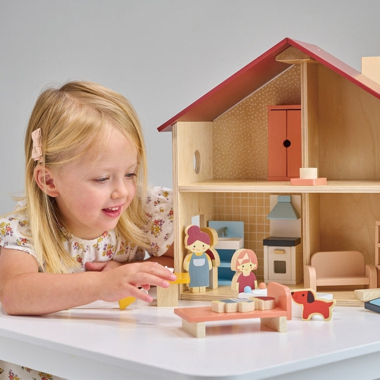 A young child playing with an open-faced wooden dollhouse by Mentari, arranged on a white table with miniature wooden furniture and small figurines.