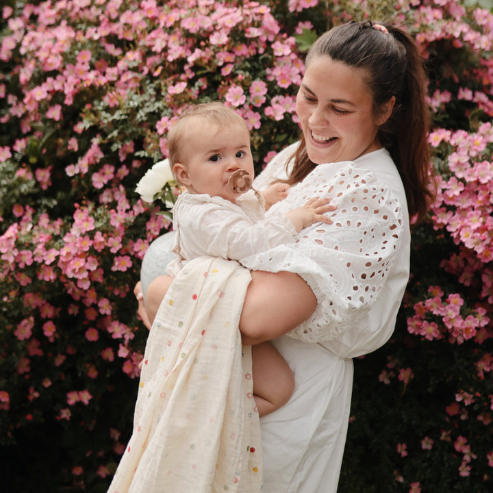 smiling woman holding a baby wrapped in a white smiley-face muslin swaddle blanket outdoors in front of pink flowers.