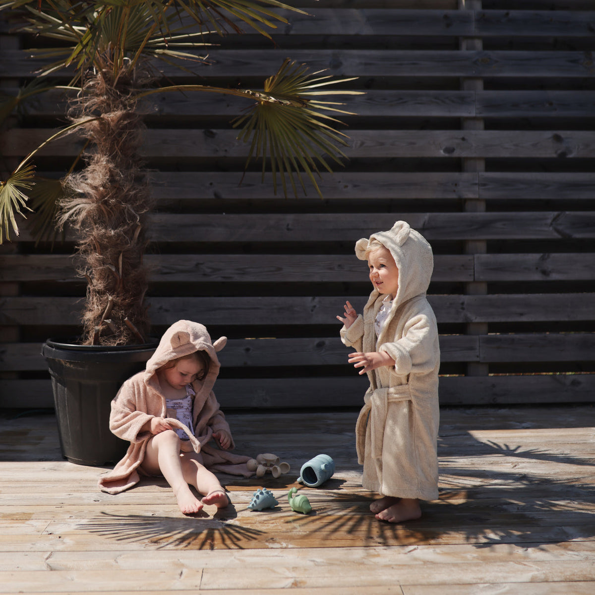 Two children in bathrobes playing outdoors on a wooden deck with a wooden wall and plant in the background.