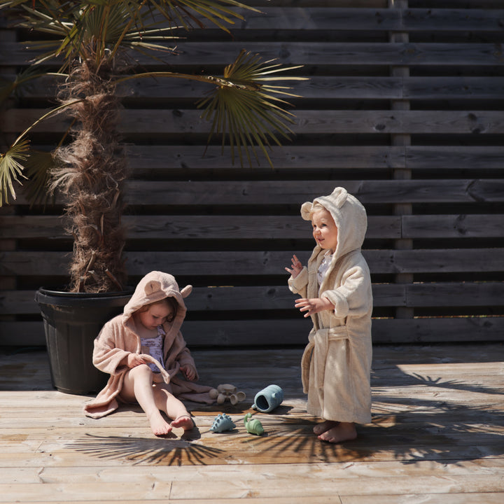 Two children in bathrobes playing outdoors on a wooden deck with a wooden wall and plant in the background.
