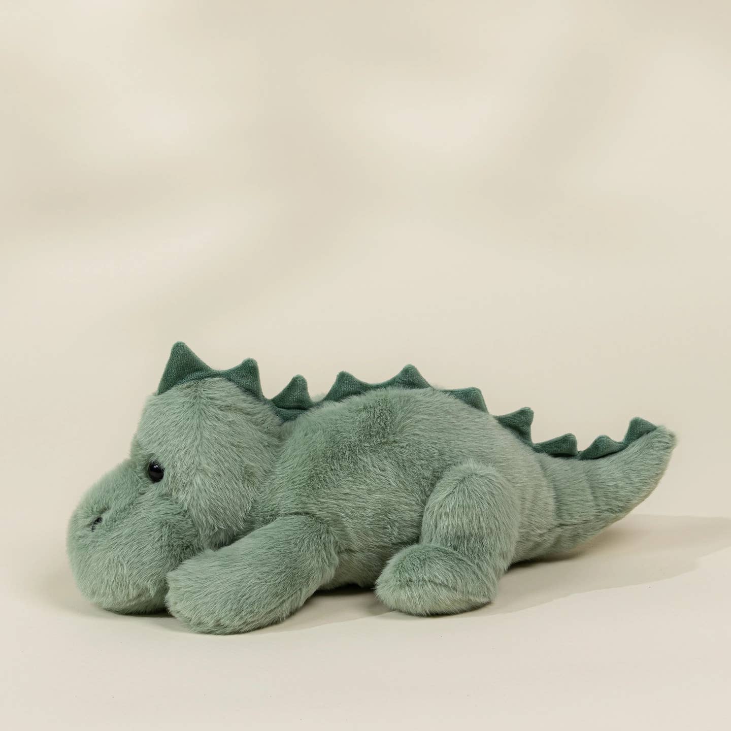 A plush dinosaur toy in a dusty sage green color, lying down on a plain light beige background.