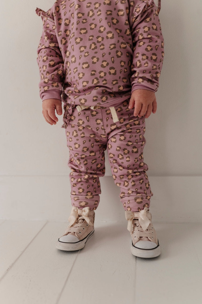 A toddler standing in a matching dusty pink leopard-print pajama set with beige sneakers, photographed against a clean white studio background.