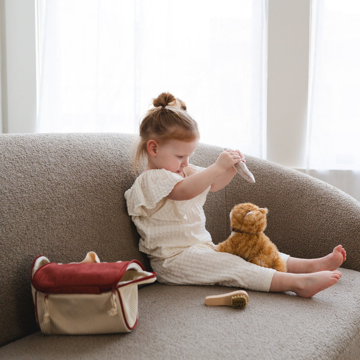 Child sitting on a couch with a teddy bear and a small bag, in a bright room.