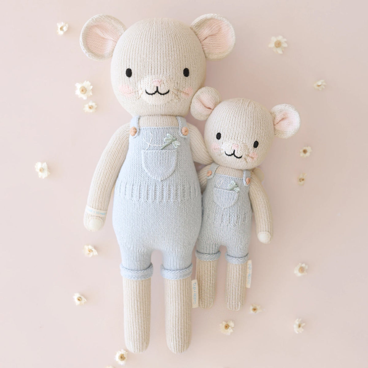 Two knitted teddy bears wearing light blue overalls on a soft pink background with small white flowers.