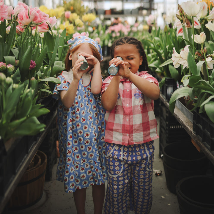 Two young children standing outdoors among blooming flowers and greenery, each holding up a Mushie kaleidoscope to one eye.