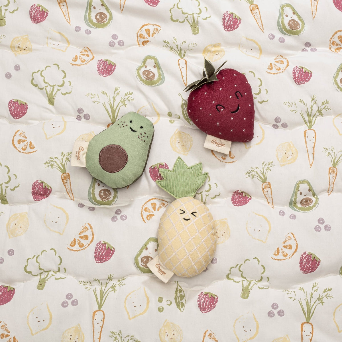 Plush toys shaped like an avocado, strawberry, and pineapple on a fabric with vegetable pattern