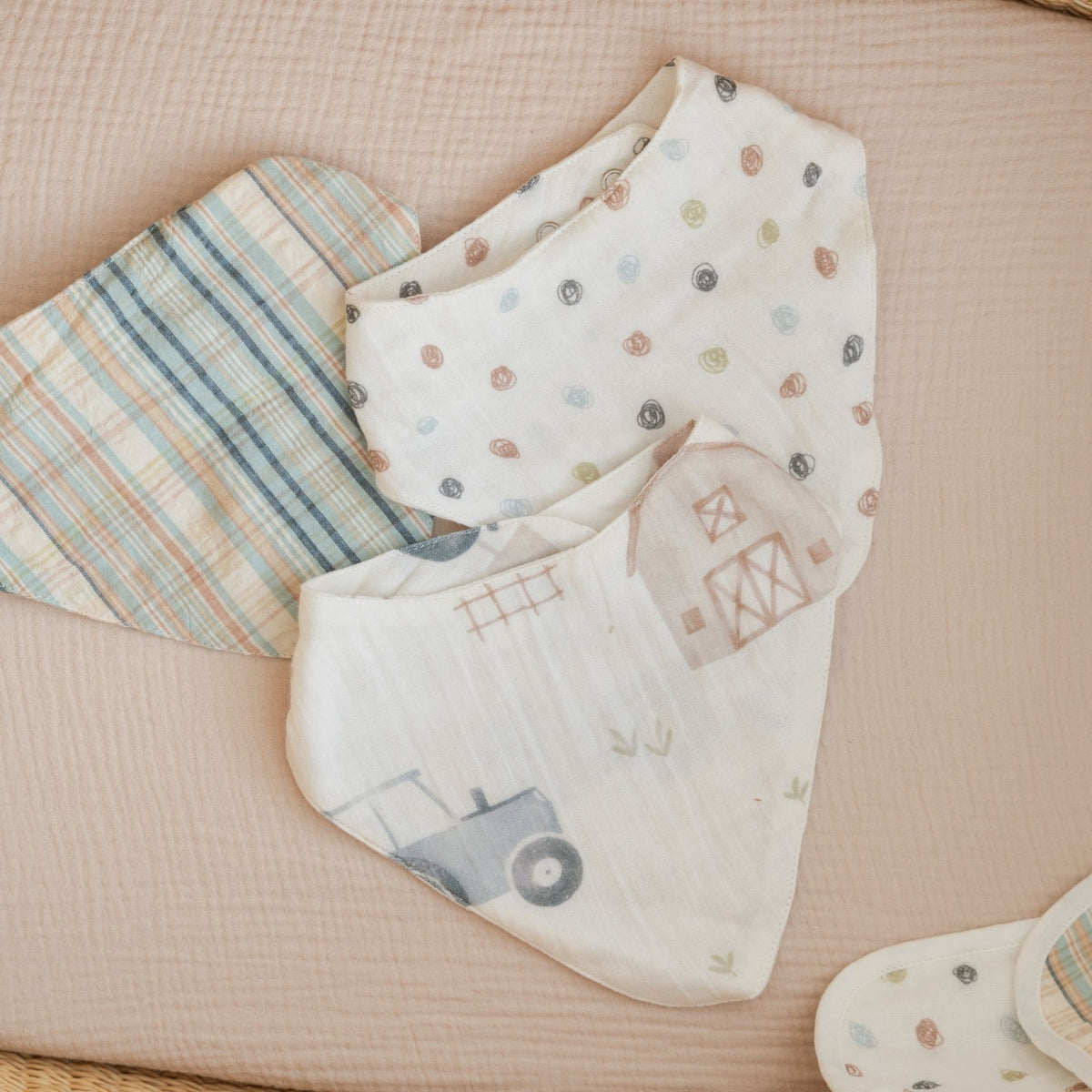 Three reusable baby feeding cloths or muslin baby bibs laid flat on a soft beige textured blanket. One cloth has a blue, green, and beige plaid pattern, while the other two are off-white with small multicolor polka dots and a barn illustration.