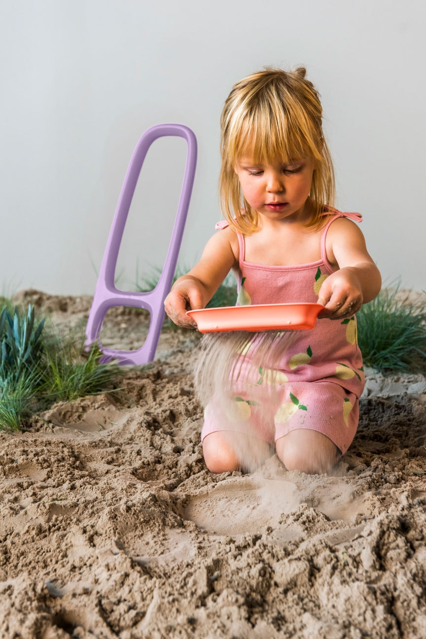 Child playing in sand with a pink shovel and orange pan on a sandy surface.