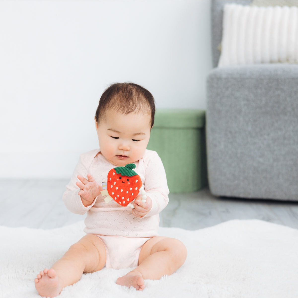 Baby sitting on a white blanket holding a toy strawberry in a minimalistic room.