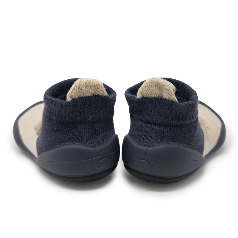 A pair of slip-on baby booties shown from the back, featuring navy blue knit socks with a beige inner lining and a matching navy blue flexible sole.