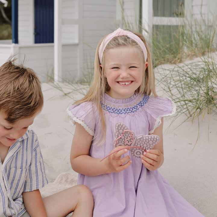 A young girl with blonde hair sits on a sandy beach next to a boy, wearing a lavender dress with blue embroidery and holding a butterfly-shaped accessory. A white beach house and dune grass are in the background.