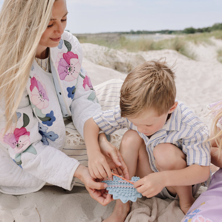 A woman and a young boy sitting on sand at a beach, both wearing matching white floral and striped pajamas while holding a small blue textured toy.