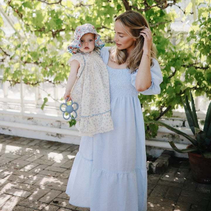 Woman in a light blue dress holding a child in a floral hat outdoors.