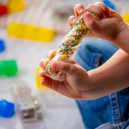 A child holding and examining a translucent Jelly Blox brick filled with colorful mini beads, with other colorful building blocks scattered in the background.