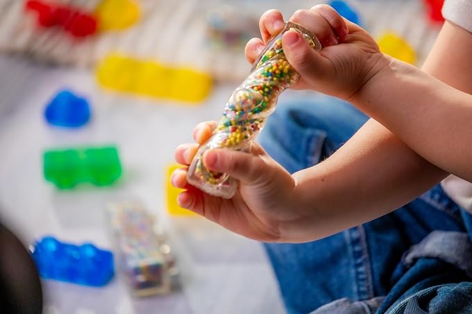 A child holding and examining a translucent Jelly Blox brick filled with colorful mini beads, with other colorful building blocks scattered in the background.
