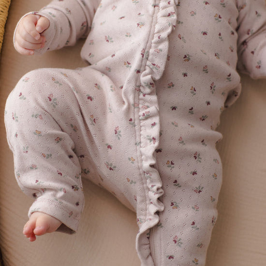 A close-up of a baby wearing a light pink long-sleeve footless sleeper with a delicate multicolor floral print and ruffle trim down the front, lying on a beige blanket.