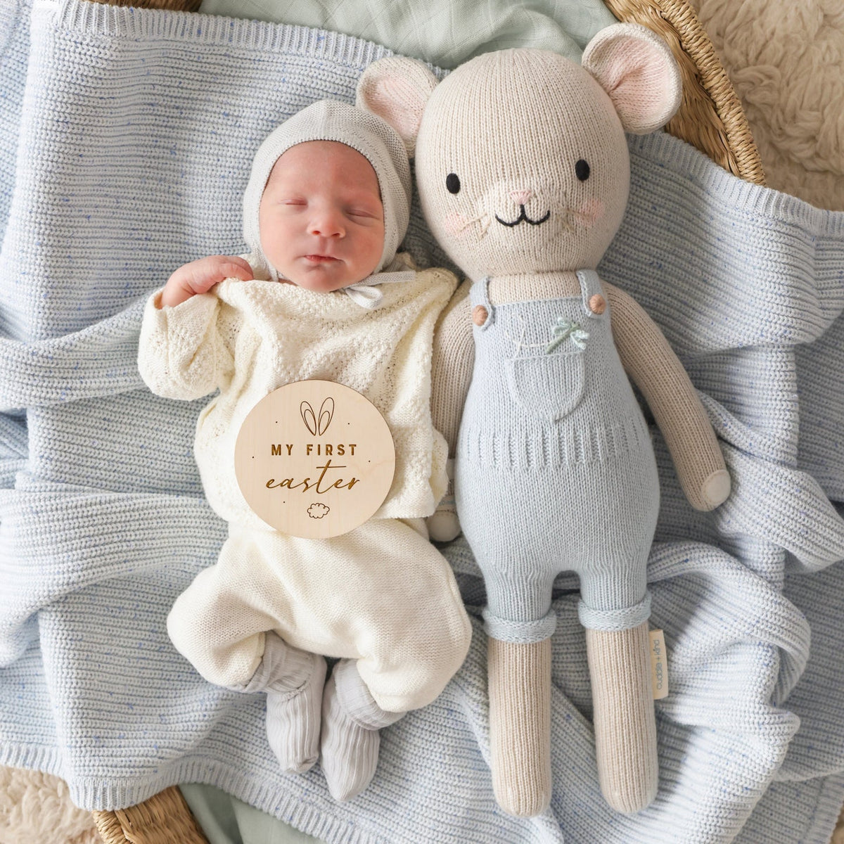 Newborn baby in a cream outfit with 'My First Easter' sign, lying next to a plush toy on a textured blanket.