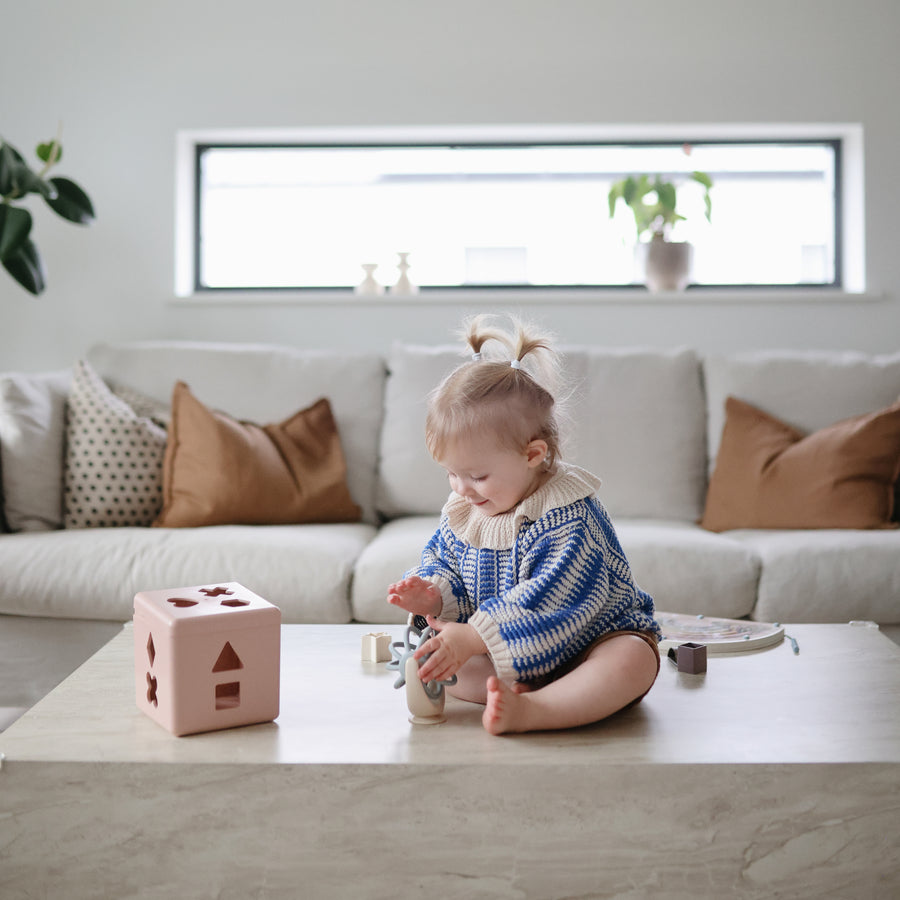 A toddler wearing a blue and white knit sweater sitting on a living room floor next to a pink toy block, playing with a Mushie Suction Spinner Ball.