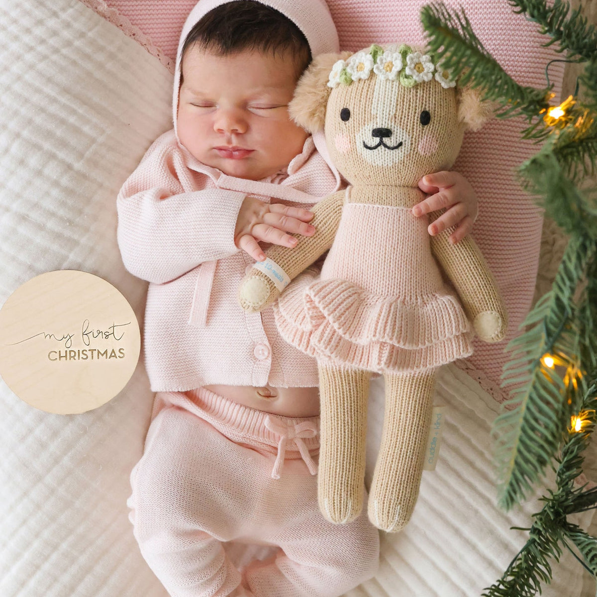 A sleeping newborn baby wearing a soft pink knit bonnet, cardigan, and footed pants, cuddling a knitted teddy bear doll with a flower crown. The baby is lying on a textured cream quilt with a pink pillow, and evergreen branches with warm string lights frame the scene.