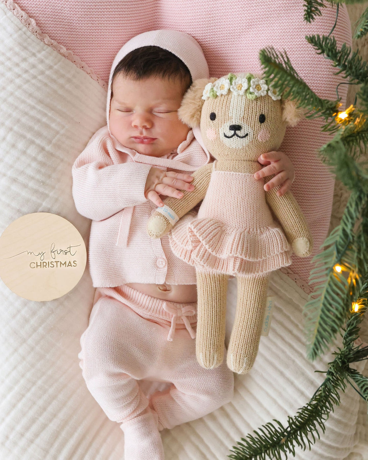 A sleeping newborn baby wearing a soft pink knit bonnet, cardigan, and footed pants, cuddling a knitted teddy bear doll with a flower crown. The baby is lying on a textured cream quilt with a pink pillow, and evergreen branches with warm string lights frame the scene.