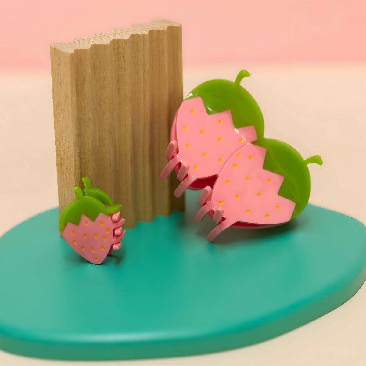 Pink strawberry-shaped hair clips on a turquoise stand with a wooden block against a pink background