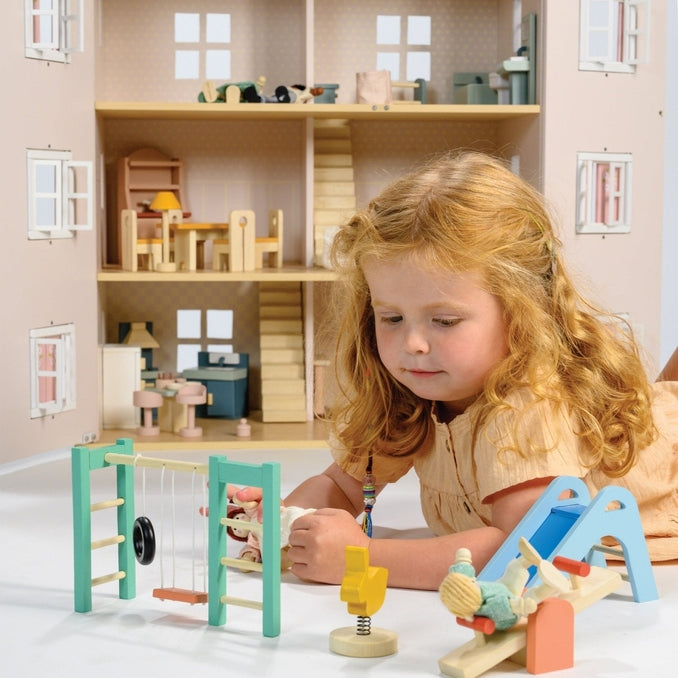 A young child playing on the floor with a large wooden dollhouse, accompanied by a wooden playground set that includes a slide, see-saw, climbing frame, swing, tire, and a bouncy bunny toy.