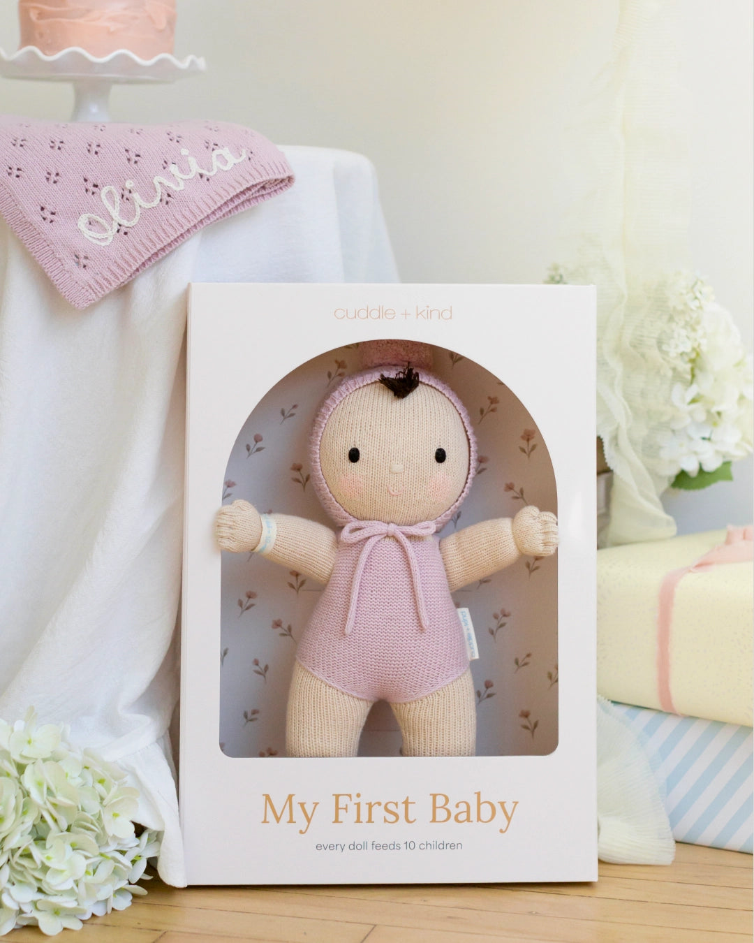 Baby doll in a pink outfit in a box labeled 'My First Baby' with a pink blanket and flowers in the background.