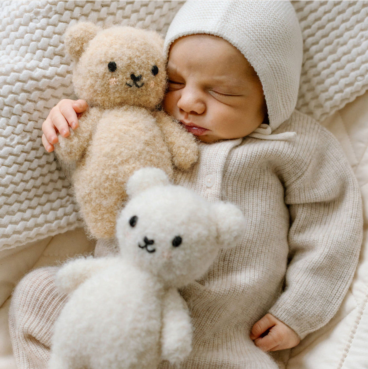Baby in a beige outfit holding two teddy bears on a textured blanket