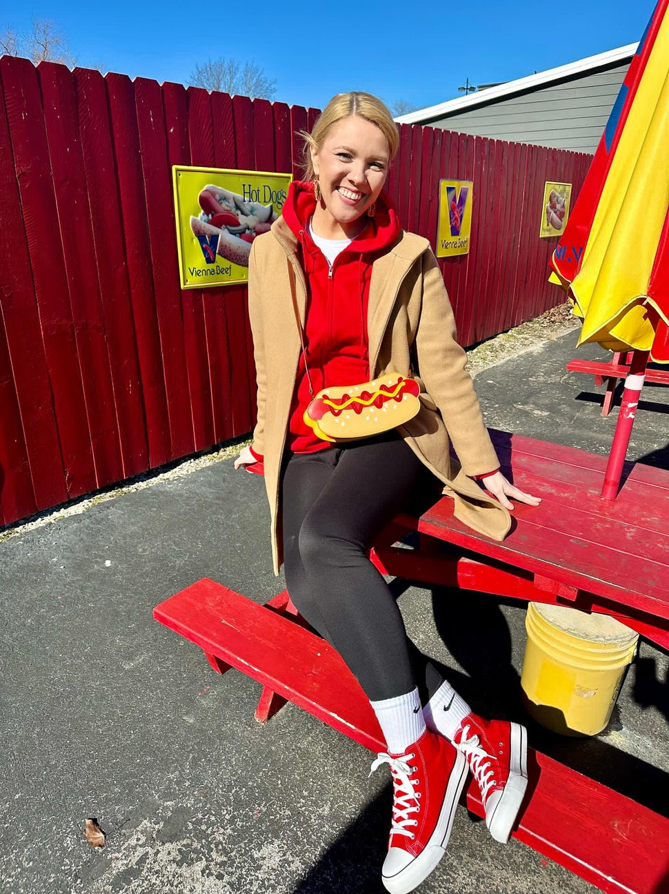 Person in a hot dog costume sitting on a red bench with a red fence and colorful decorations in the background.