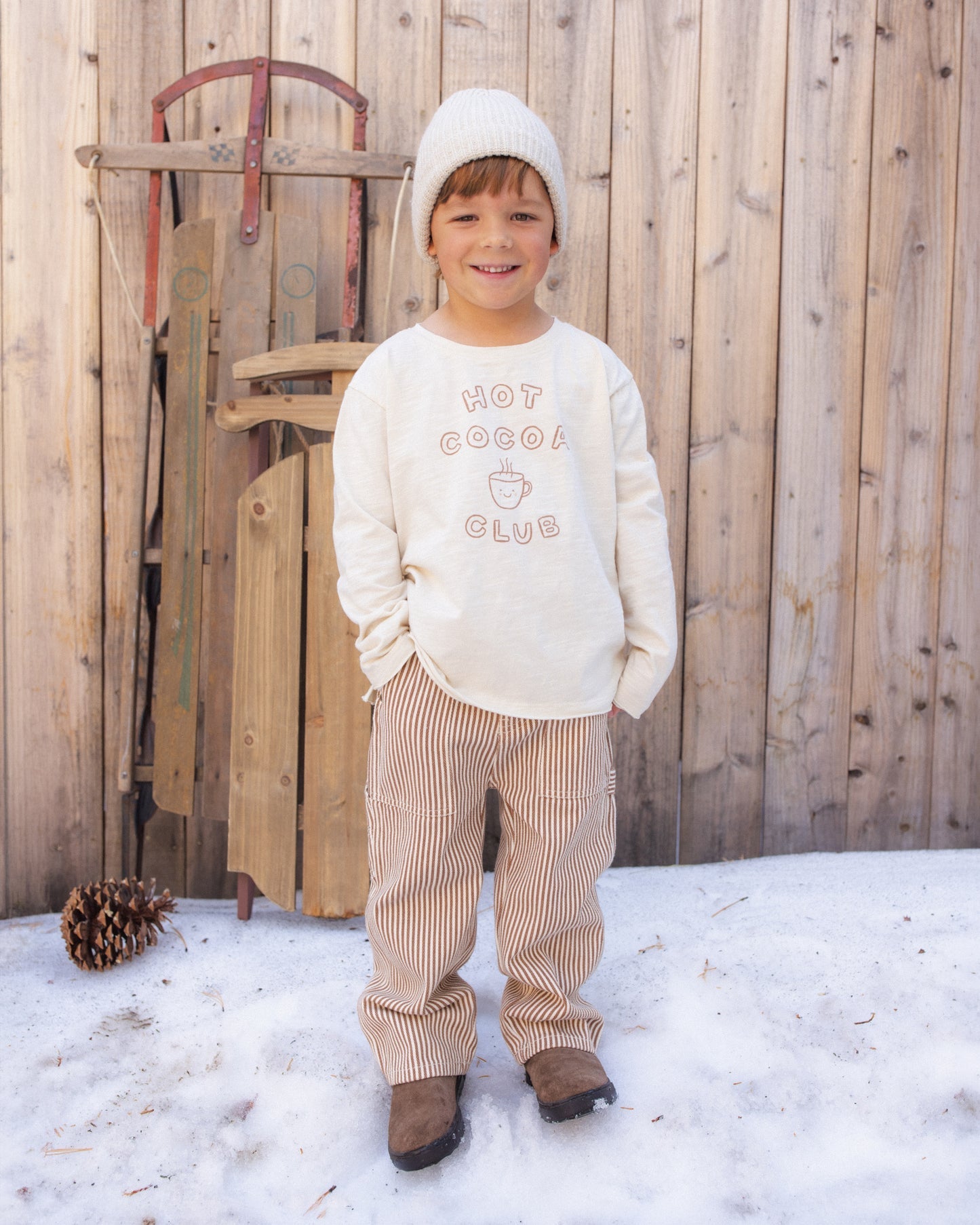 A young child standing outdoors in front of a wooden fence and snow, wearing a cream sweatshirt with 'HOT COCOA CLUB' text, cream knit beanie, tan pinstriped pants, and brown boots.