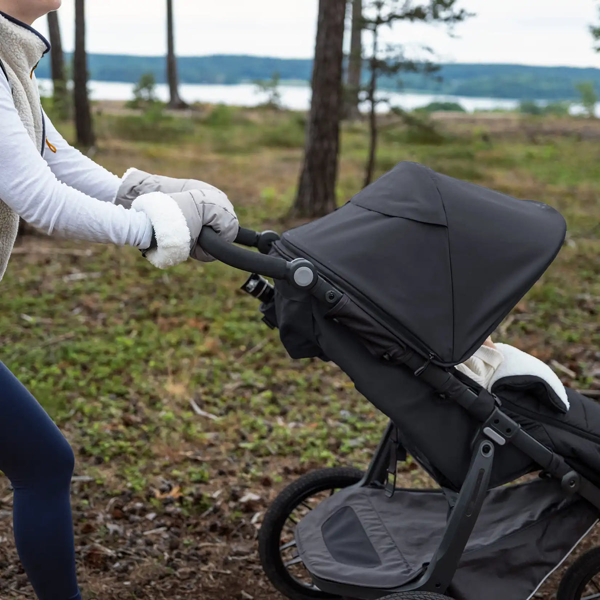 Person pushing a black stroller in a natural setting with trees and water in the background