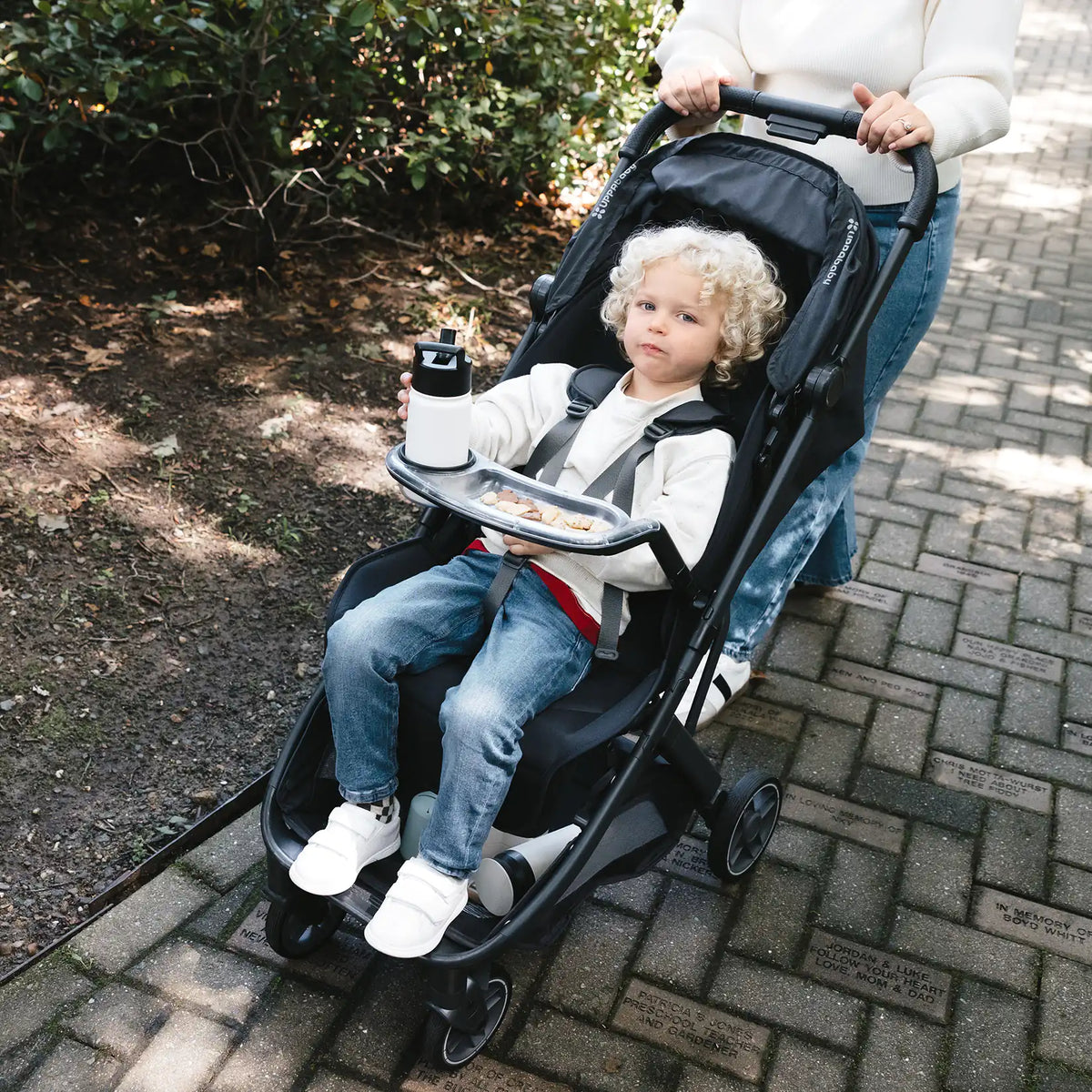 A young toddler with curly blonde hair sits comfortably in an UPPAbaby Minu V3 stroller outdoors on a paved stone pathway, holding a drink bottle on the attached snack tray.