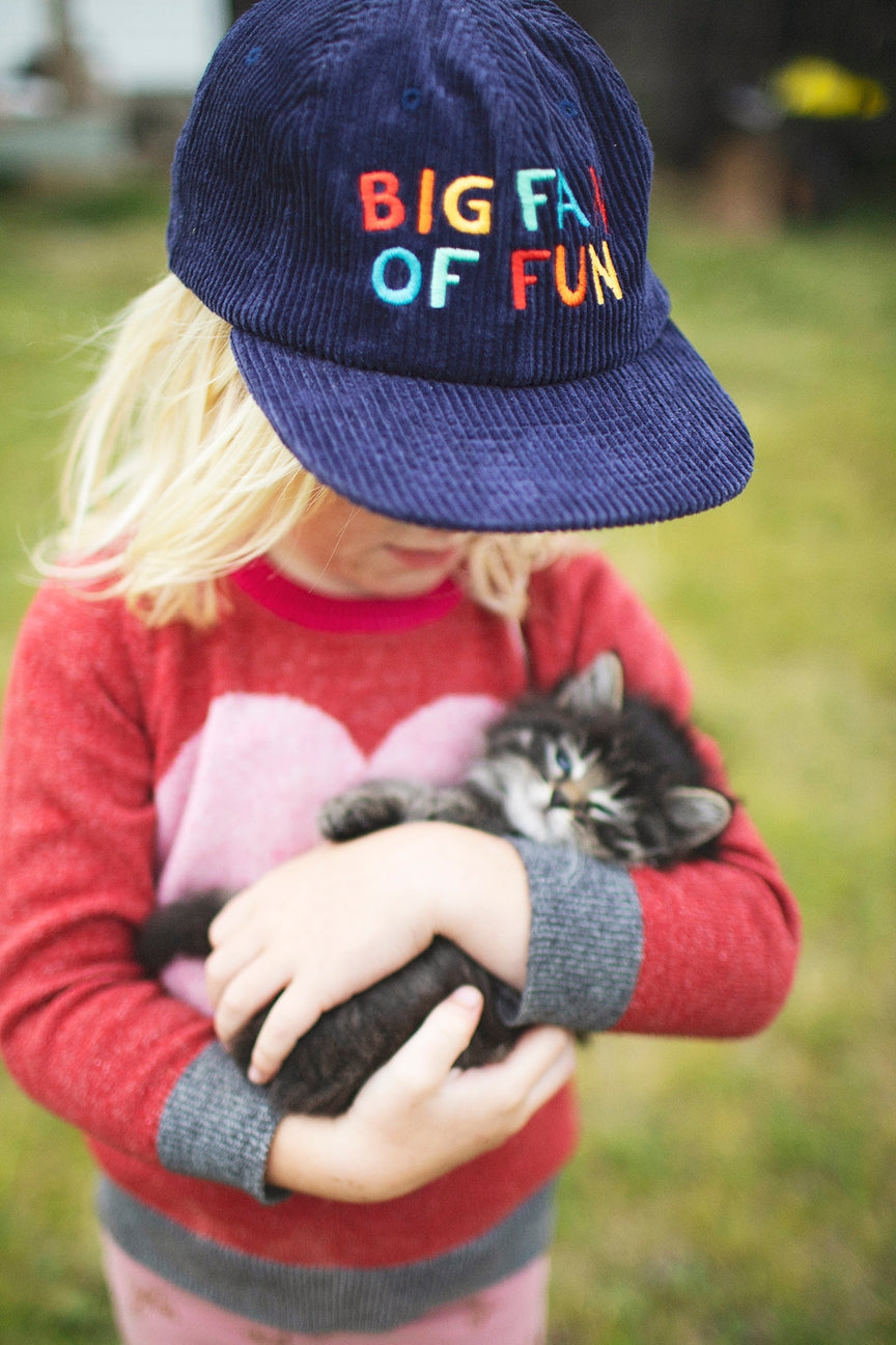 Child wearing a navy cap with colorful text, holding a kitten, outdoors.