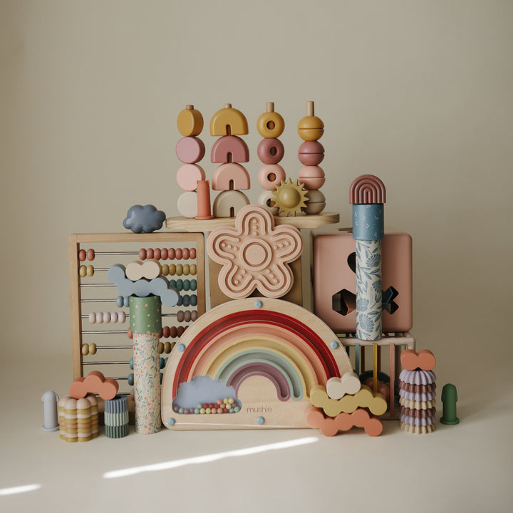 A flat lay arrangement on a white background featuring two Mushie kaleidoscopes surrounded by wooden toys, silicone stackers, animal figurines, an abacus, and space-themed pieces.