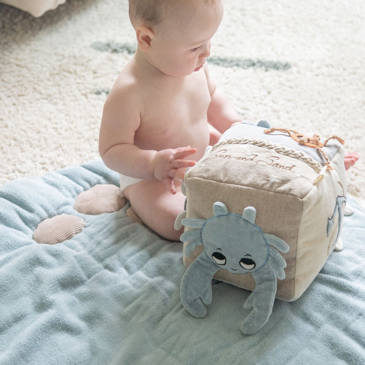 A baby sitting on a light blue blanket next to a beige cube-shaped plush toy with a blue octopus face and embroidered text.