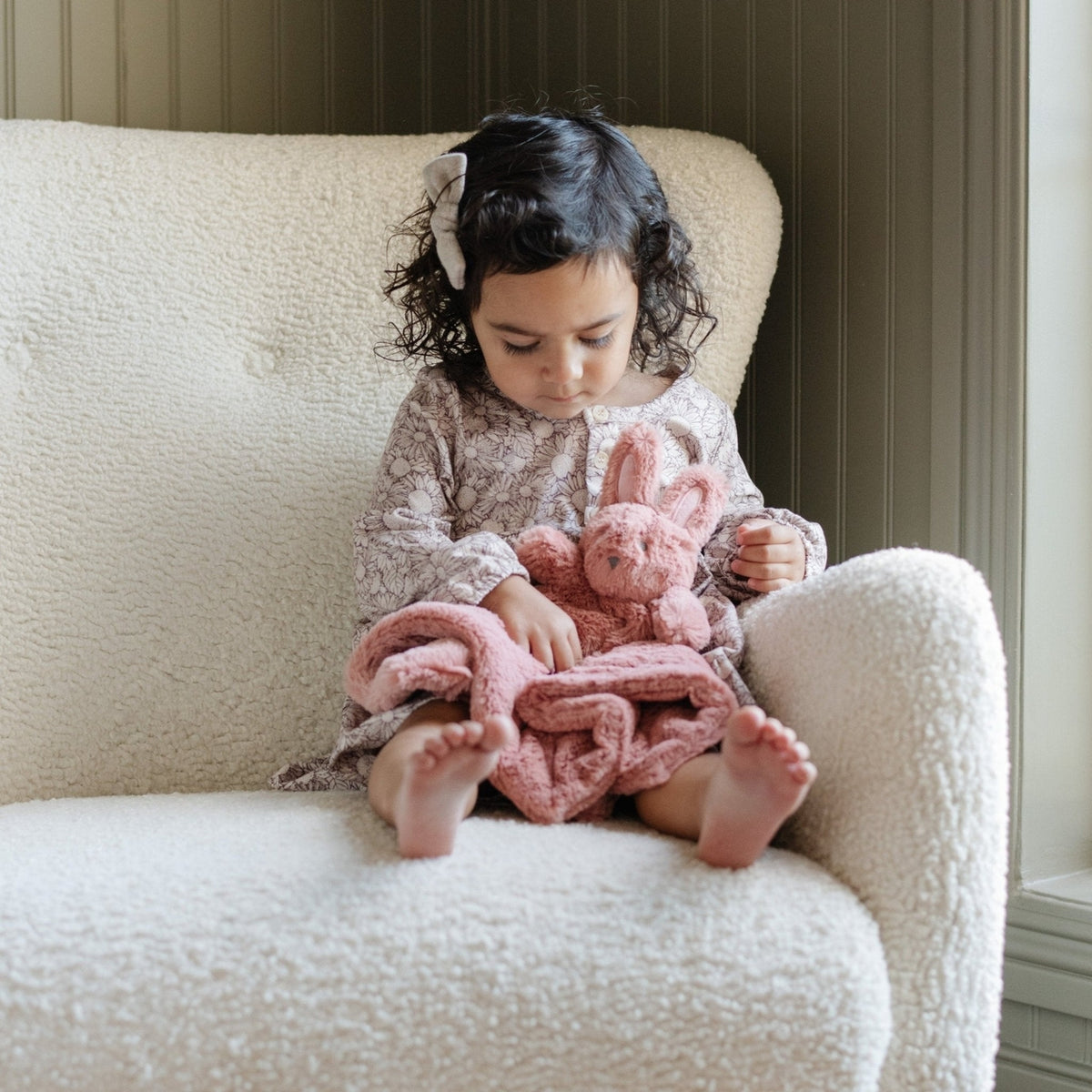 Child sitting on a white chair holding a pink knitted blanket and toy
