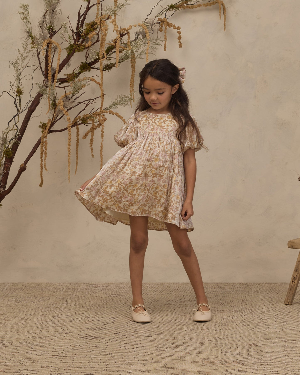 A young girl with long dark hair stands in a studio wearing a short cream dress with a yellow floral print and flutter sleeves. She is posed in front of a textured beige wall with decorative dried branches on the left.