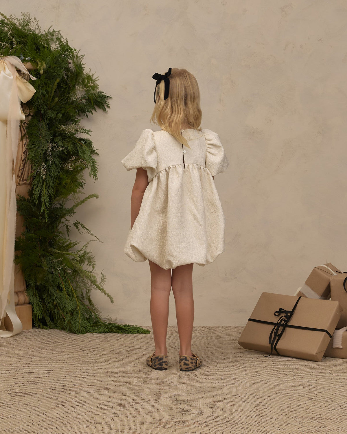A young girl with blonde hair tied back with a black bow is shown from behind wearing a short cream-colored puff-sleeve dress. She is standing in a styled studio setting with a textured beige wall, a green foliage arrangement on the left, and wrapped gift boxes on the floor to the right.