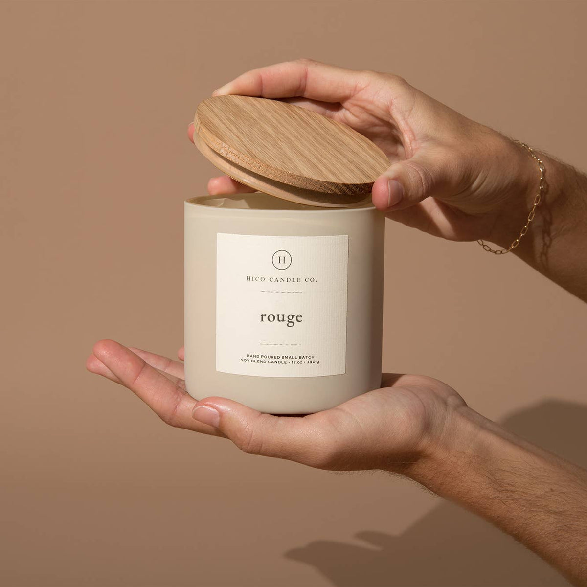 Hand holding a beige candle labeled 'rouge' with a wooden lid against a brown background
