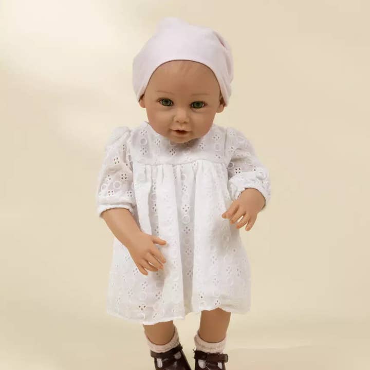 A realistic baby doll with a white eyelet dress, white bonnet, and dark brown shoes, standing against a plain beige background.