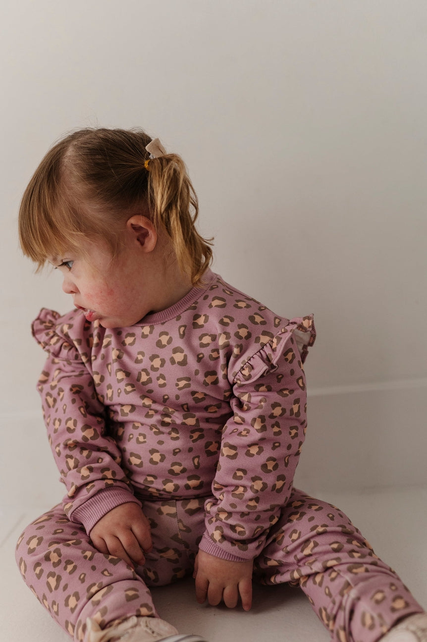 A toddler girl sitting on a white floor against a white wall, wearing a dusty purple long-sleeve pajama set with a small yellow and white floral print.